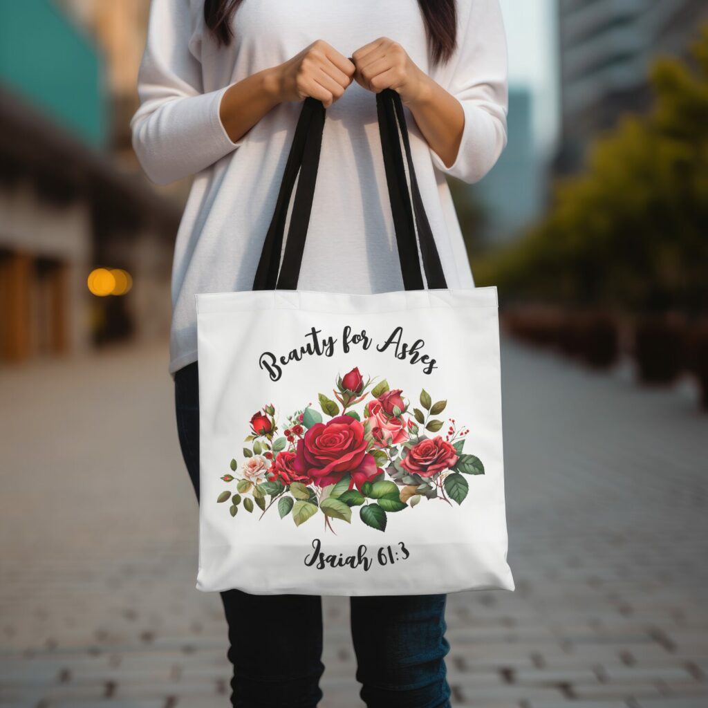 this image shows a woman's hands holding a tote bag with a bouquet of beautiful roses on it and the words 'beauty for ashes, isaiah 61:3' it shows the design of a beautiful bag for anyone hurting and going through pain.