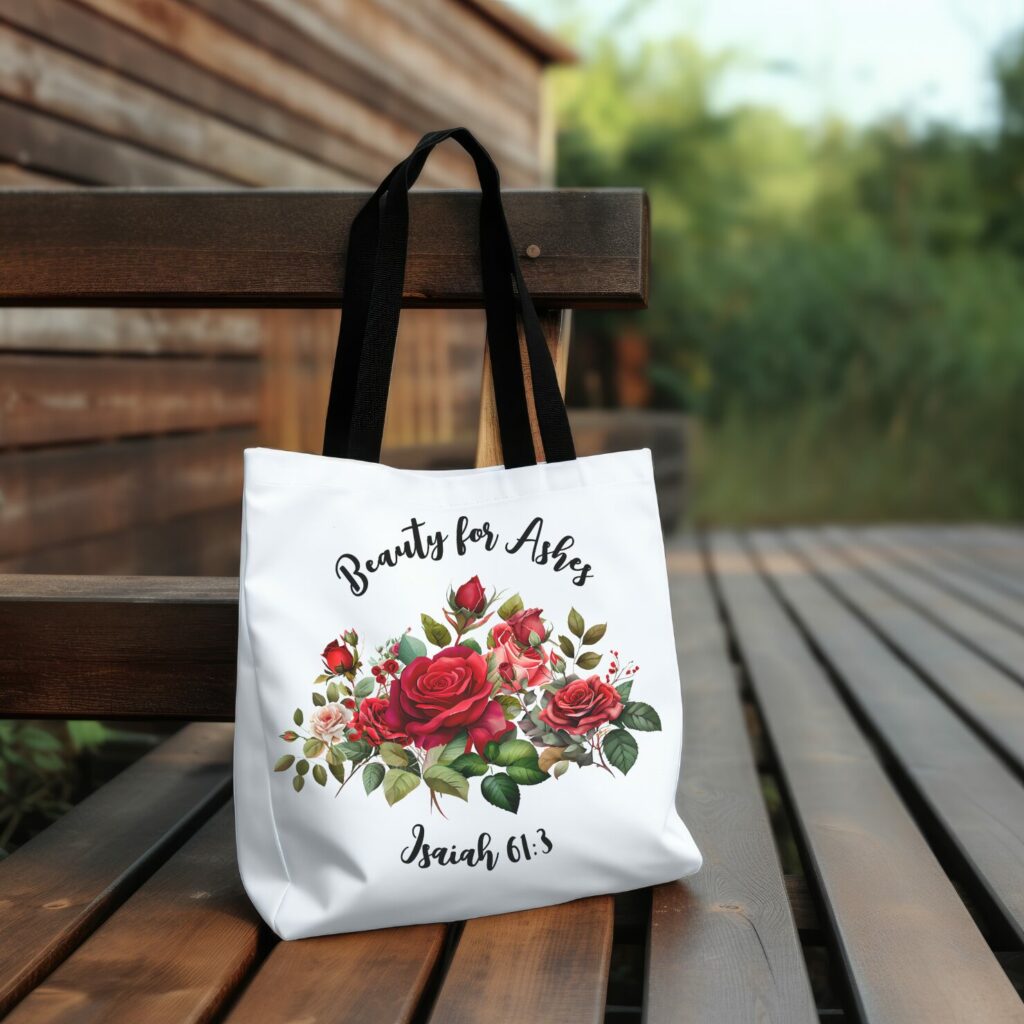 this picture shows a white tote bag with a rose bouquet on it and the words "beauty for ashes, isaiah 61:3" written across the top and bottom of the tote bag. It has a black handle on the bag and is resting on a wooden bench. In the background there is some lush greenery.